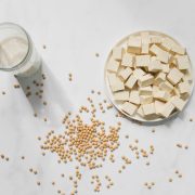 Cubos de tofu, grãos de soja e um copo de leite de soja vistos de cima sobre fundo branco.
