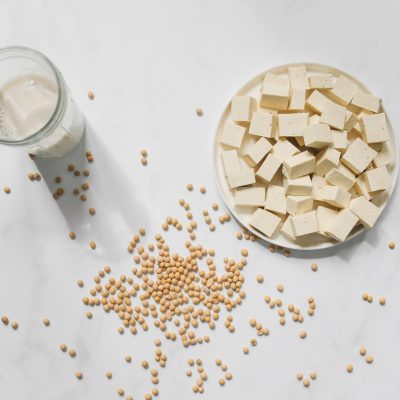 Cubos de tofu, grãos de soja e um copo de leite de soja vistos de cima sobre fundo branco.
