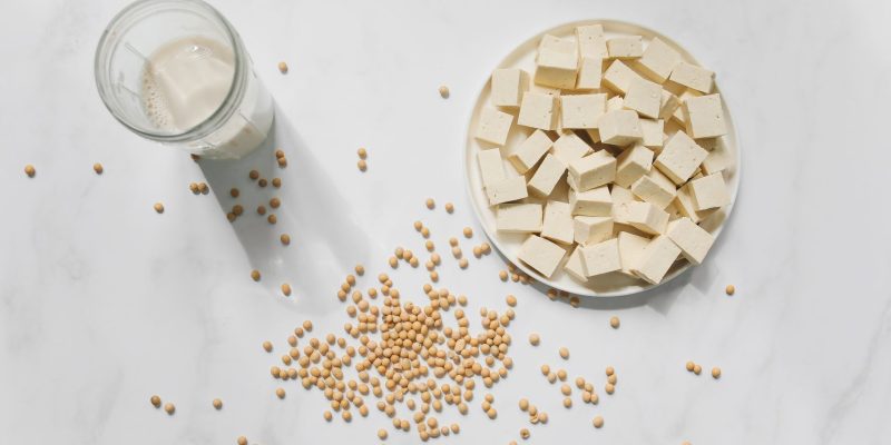 Cubos de tofu, grãos de soja e um copo de leite de soja vistos de cima sobre fundo branco.